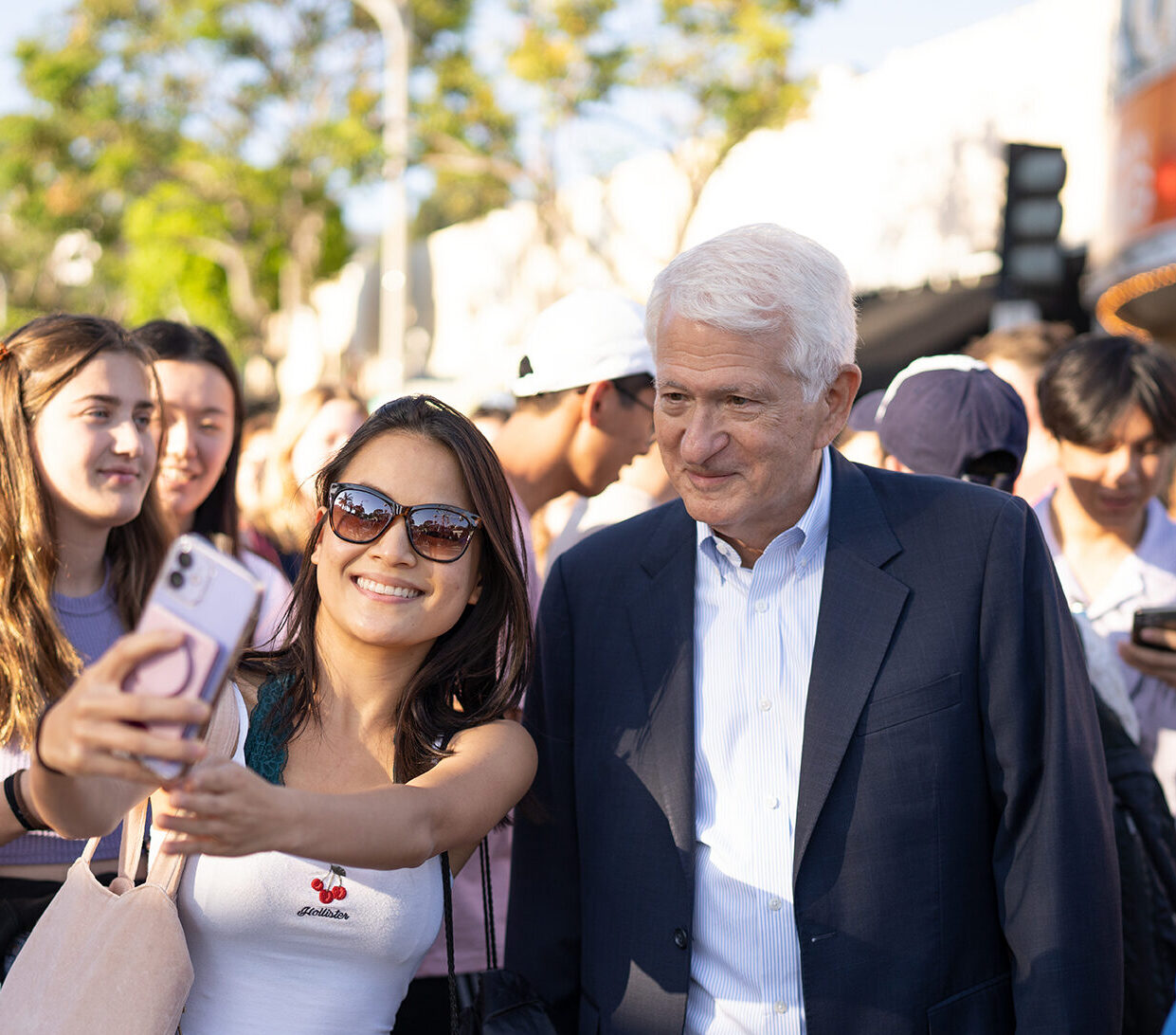 A female student in sunglasses holds her phone out to take a selfie with Chancellor Block in a crowd.