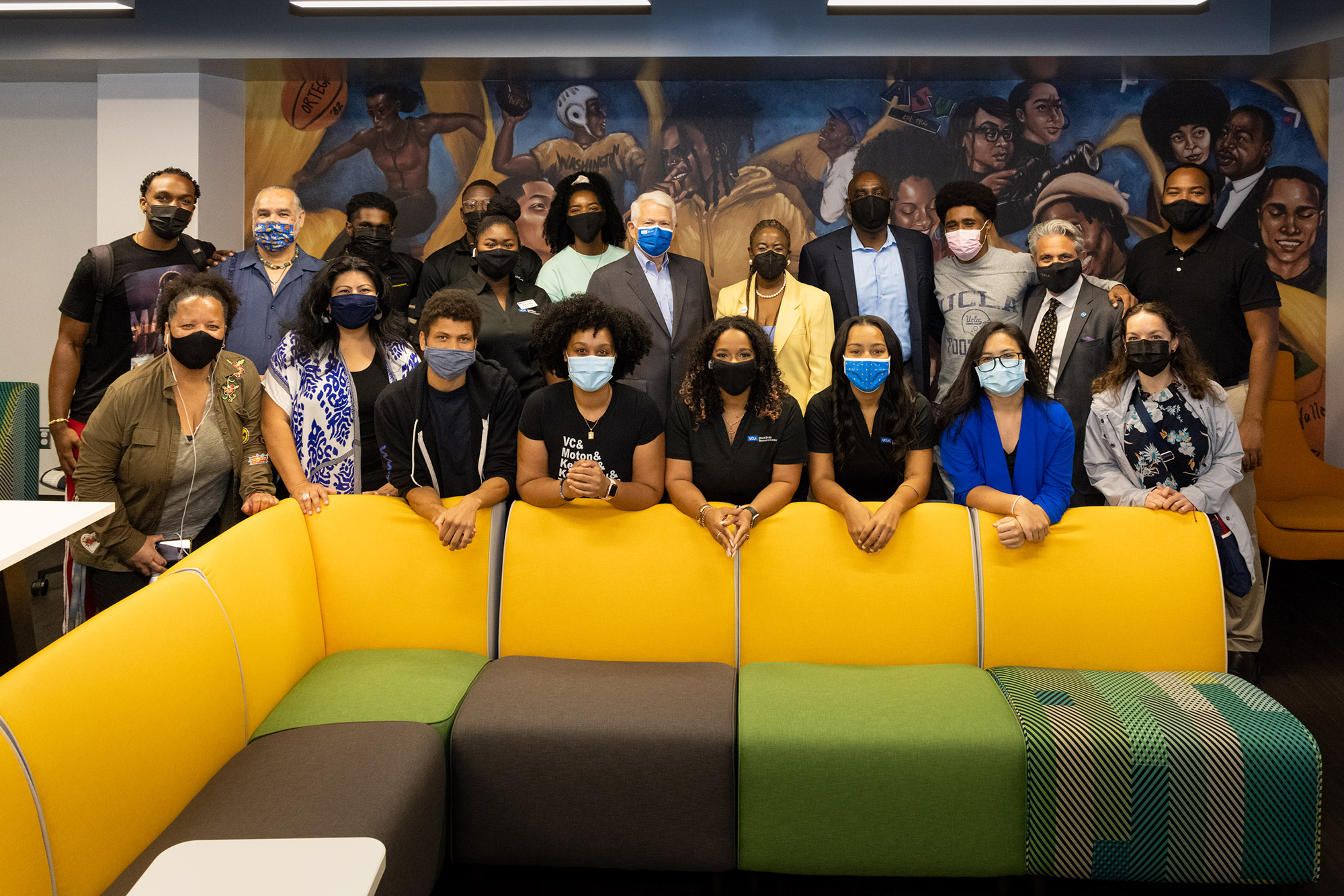 A large group donning COVID masks take space in front of a hand painted mural, depicting notable Black Bruins. Chancellor Block can be seen in the center