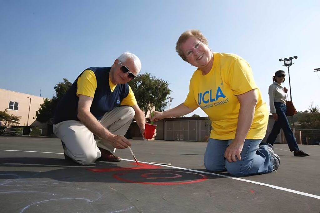 Carol Block takes a paintbrush from Chancellor Block's cup, as they kneel on the asphalt and paint a bright red letter 'L' during UCLA Volunteer Day.