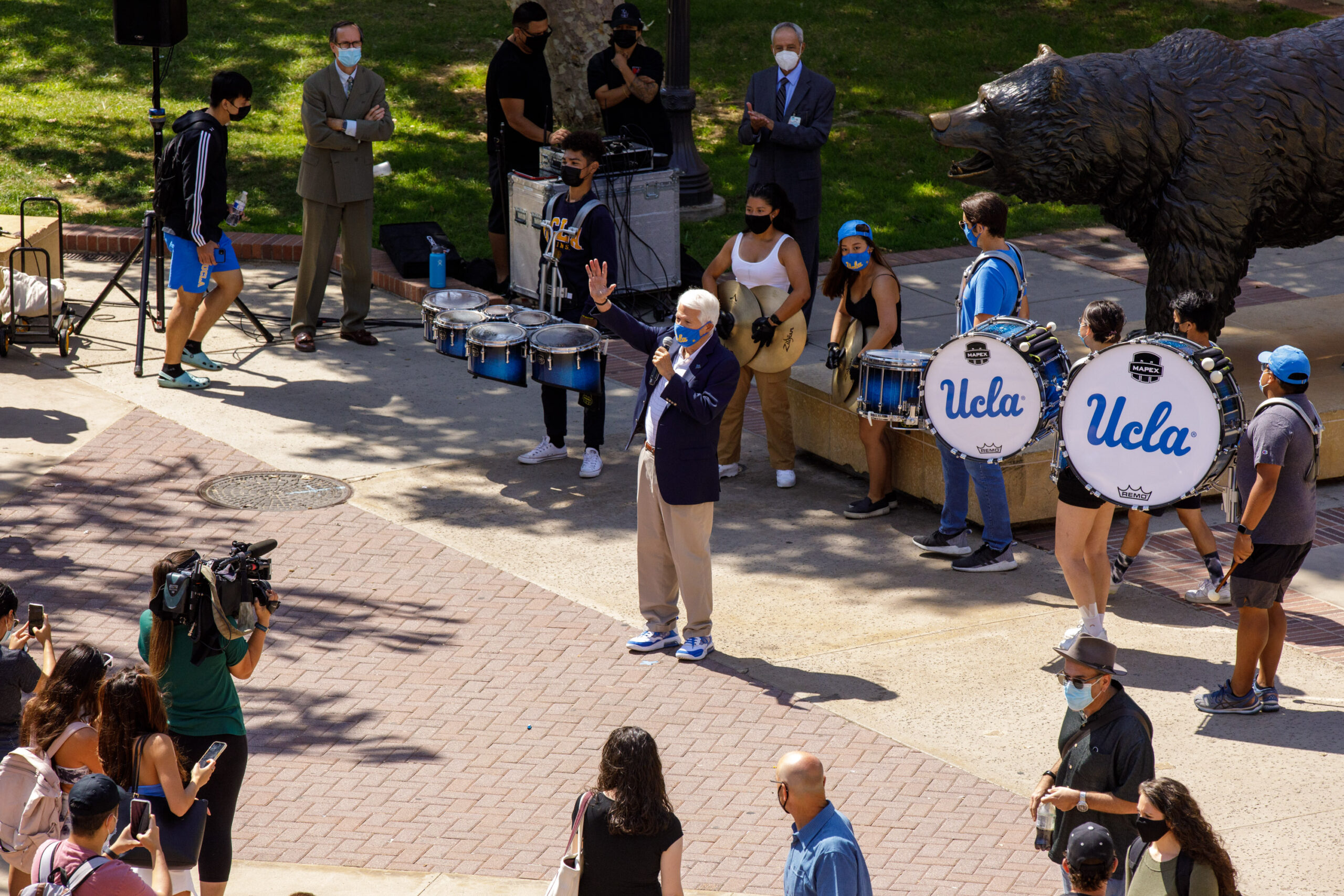 A masked grey haired man holds a microphone and raises his hand. A small crowd point cameras towards him. Behind Block, UCLA drummers and staff wrap around the Bruin statue.