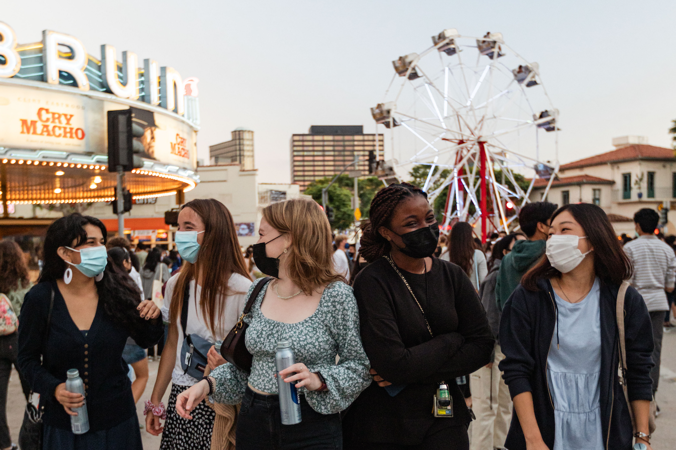 A group of masked female students laughing in downtown Westwood. Behind them a ferris wheel sits in frame.