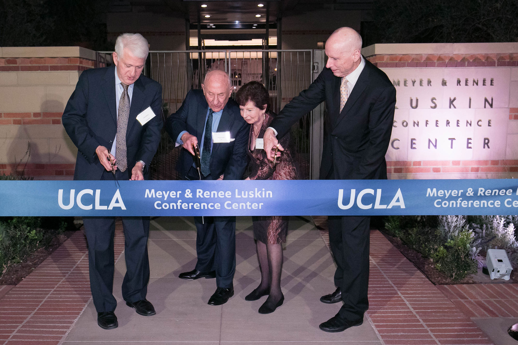A ribbon across the screen reads 'UCLA Meyer & Renee Luskin Conference Center'. Block, the Luskins, and EVCP Waugh each hold scissors to demonstrate the first snip. Background: the conference building is partially visible. 
