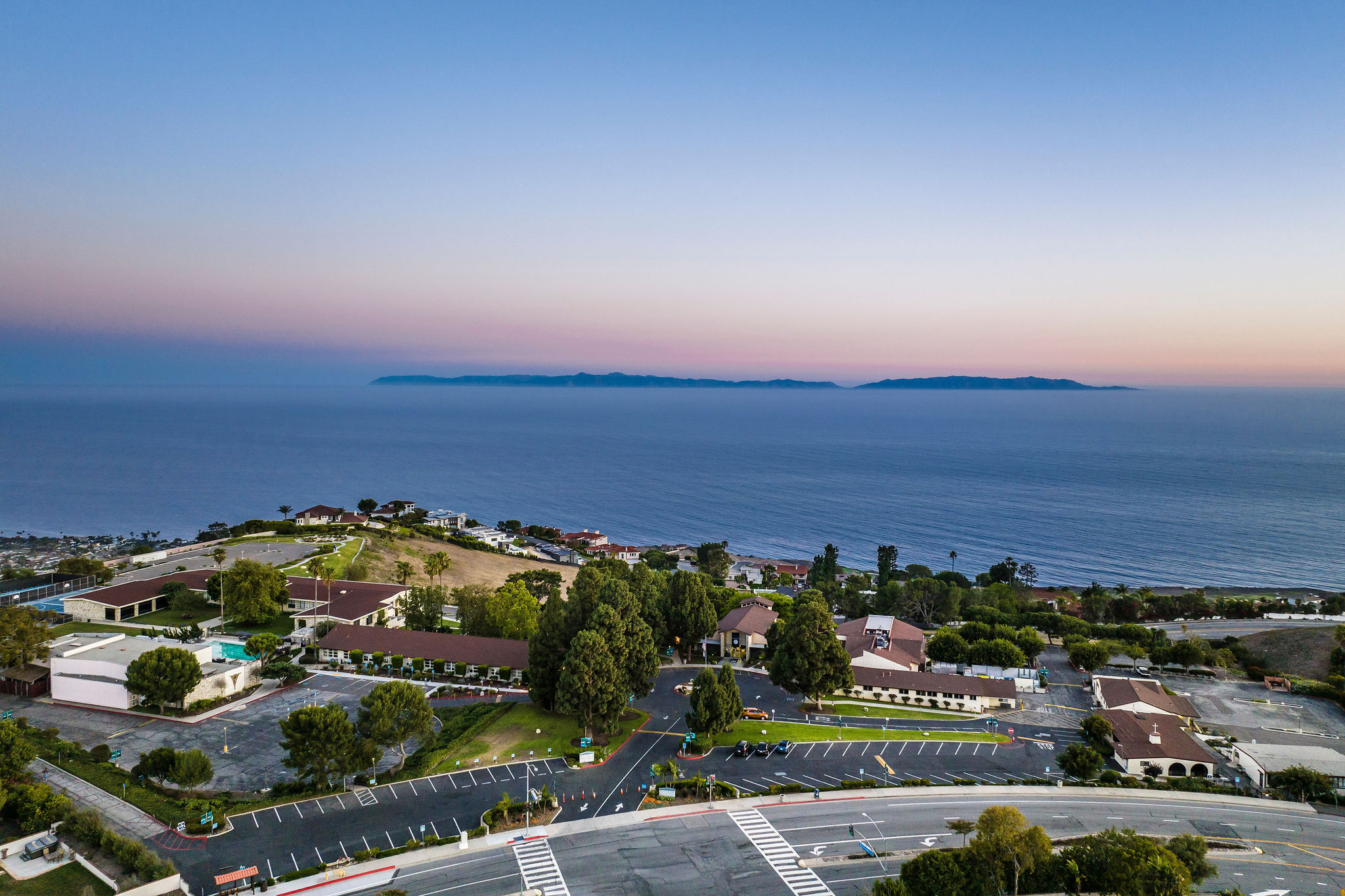 An aerial view of a parking lot lined with trees and reddish roofs, looks over the ocean as the sun sets. 