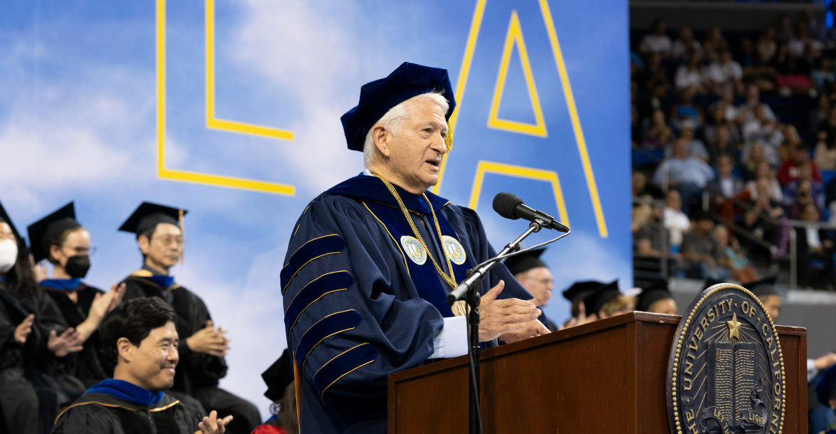 Chancellor Block in full regalia is framed by giant letters L and A. He speaks into a microphone at 2023 UCLA Commencement. Behind, Randall Park, and a large crowd clap.